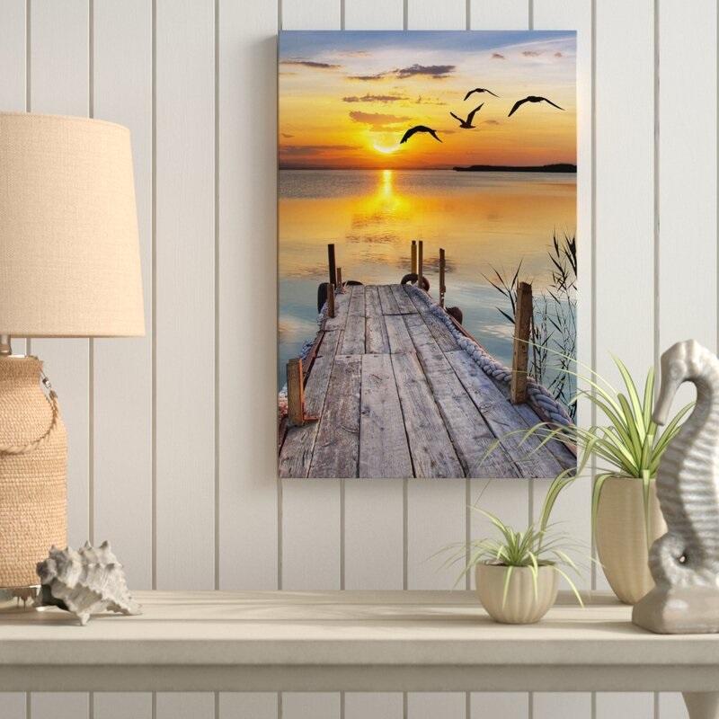 Fotografía de un muelle de madera al amanecer con aves volando y sol reflejado en el agua