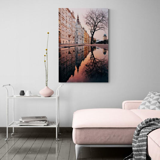 Fotografía de Praga con reflejos de edificios en charcos de lluvia y árbol deshojado en primer plano.