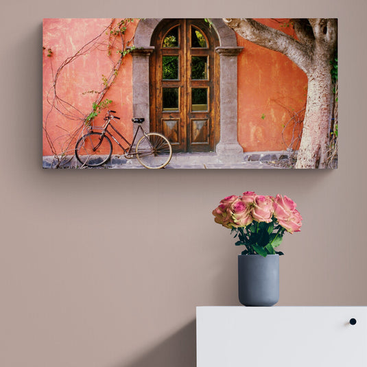 Cuadro decorativo en lienzo con diseño de bicicleta vintage apoyada en una puerta de madera, sobre una fachada color terracota con enredaderas y árbol. Estilo bohemio y nostálgico, ideal para decoración de sala o recibidor.