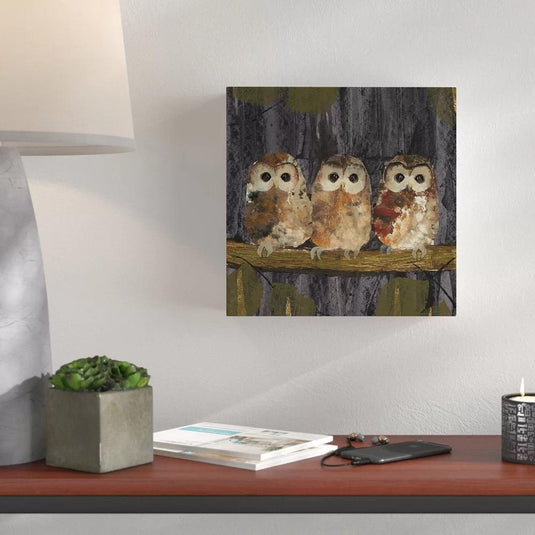 Tres búhos en tonos crema, naranja, cafés y rojo sobre rama, con fondo gris y toques verdes en cuadro decorativo