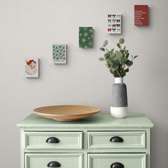 Lienzos navideños y decoración minimalista sobre cómoda verde vintage.