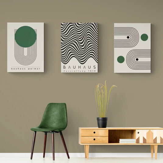 Tres cuadros de estilo Bauhaus con diseños geométricos en una pared verde oliva, acompañados por una silla moderna verde y una mesa de madera.