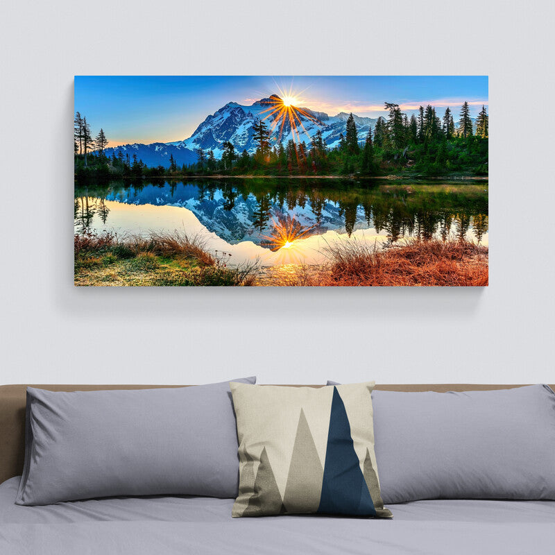 Fotografía artística de un gran lago reflejando montañas y pinos bajo un amanecer con sol emergente y cielo azul sereno